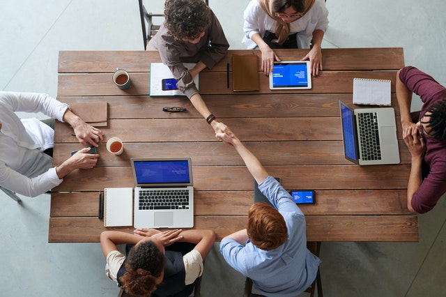 A group of six people sitting around a table with their laptops. Two of them shake hands.